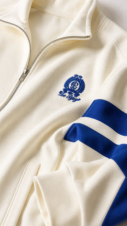 Close-up detail of cream track jacket zipper, embroidered crest, and blue sleeve stripes